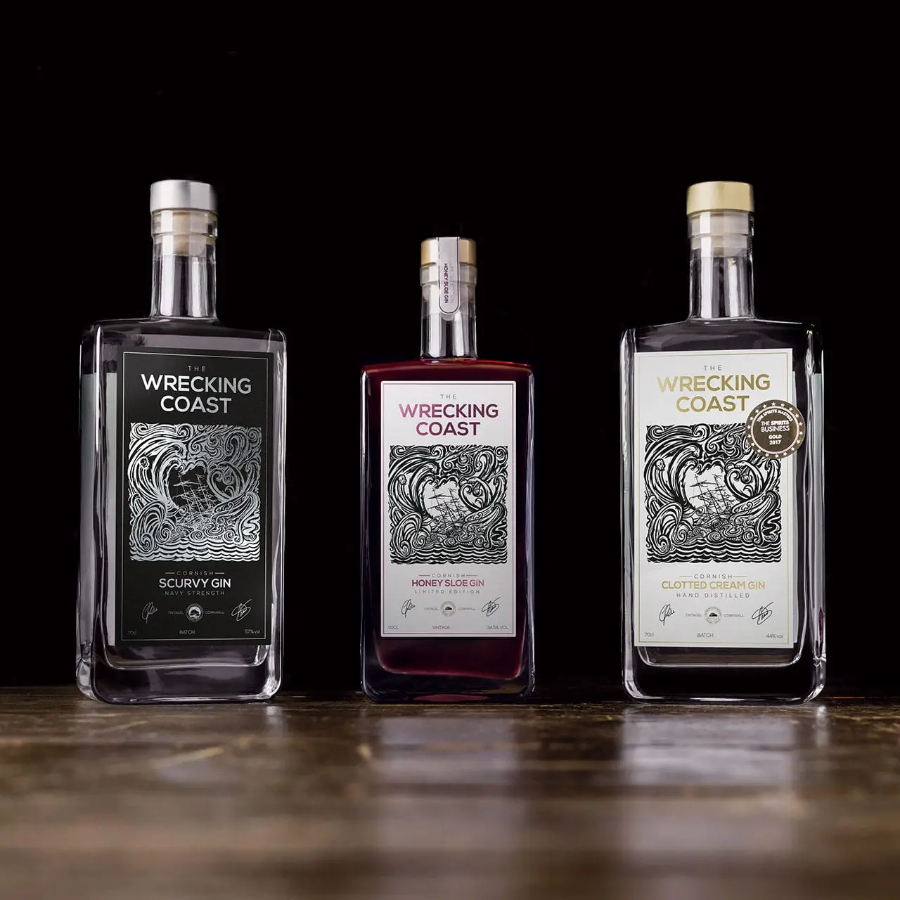 The Wrecking Coast Distillery Product Range 1280x1280 The Wrecking Coast Distillery Scurvy gin, Cornish Clotted Cream gin and Honey Sloe gin bottles, photographed on a wooden surface with a black background